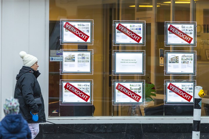 Voetganger loopt langs een makelaar met in de etalage borden van huizen die te koop en verkocht zijn.
